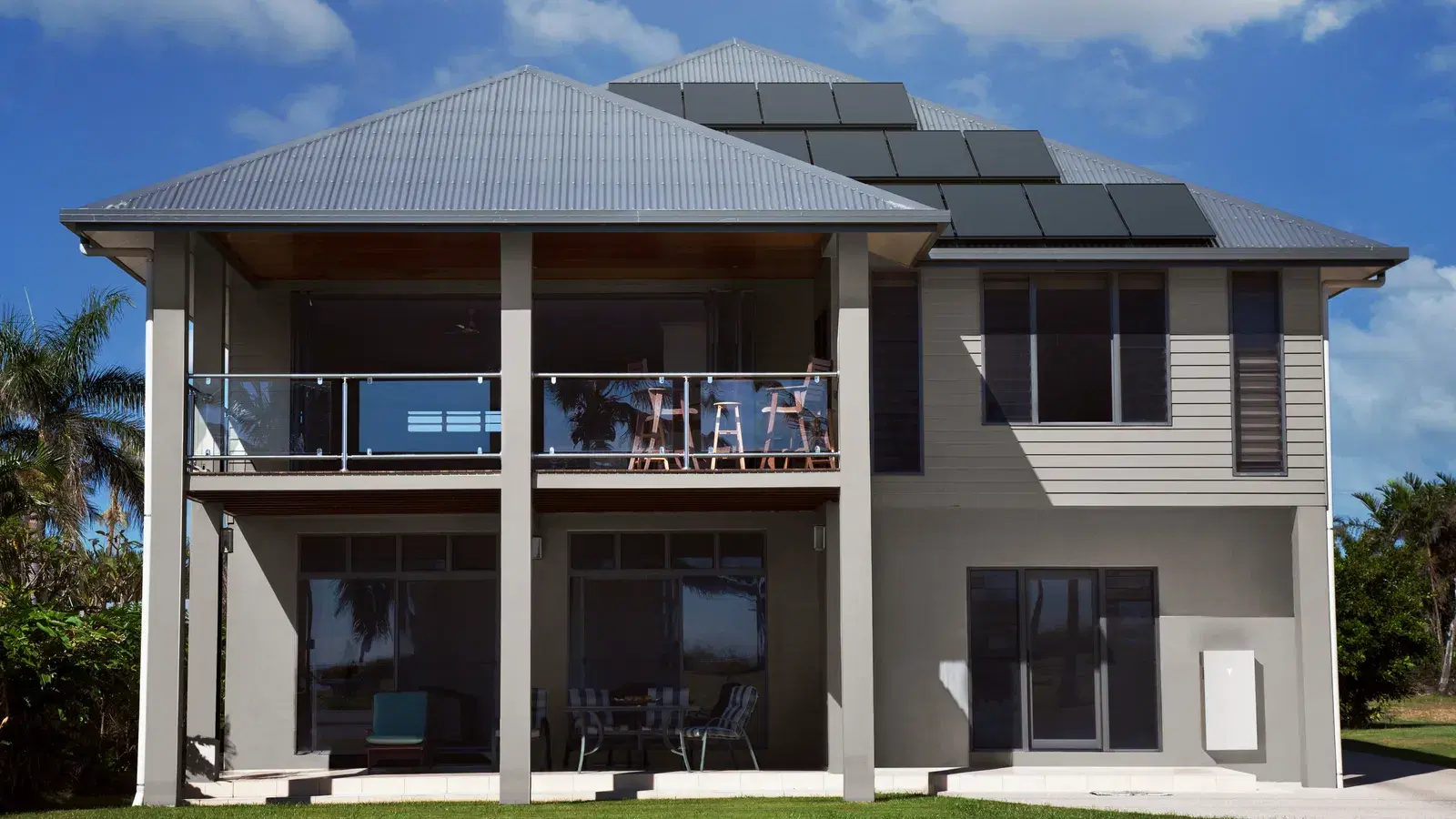 A modern gray two-story house with dark solar panels mounted on its metal hip roof against a blue sky.