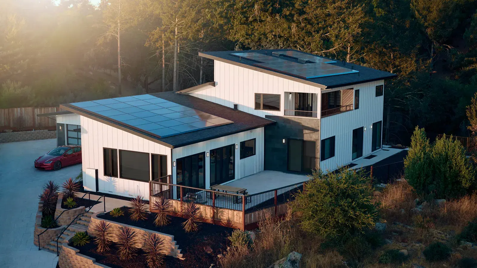 Aerial view of a contemporary white hillside home with rooftop solar panels and a red Tesla parked in the driveway at sunset.