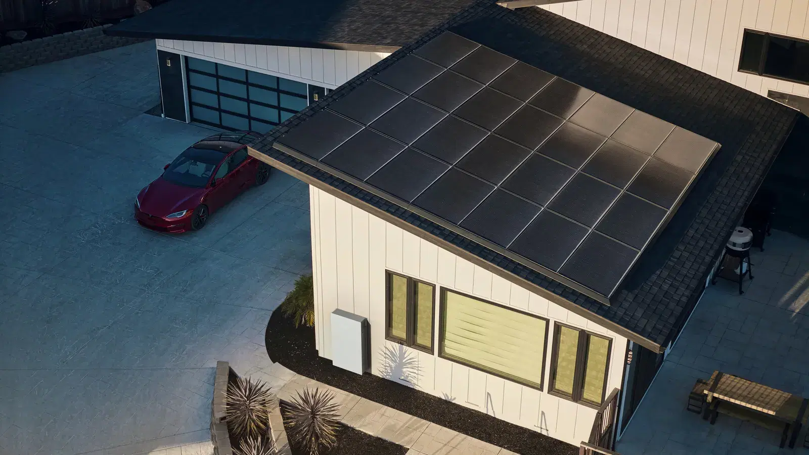 Elevated drone view of a black rectangular solar panel array on a home's roof, with a Powerwall on the wall and a car in the driveway below.