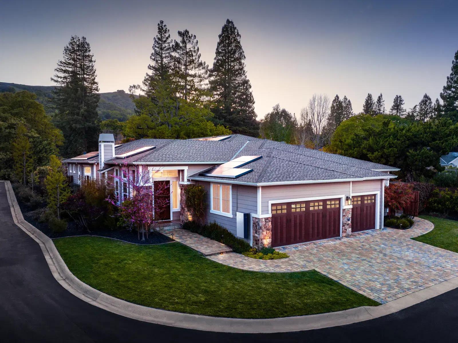 Aerial twilight view of an upscale suburban home with black solar panels across its roof, stone facade and a paver driveway.