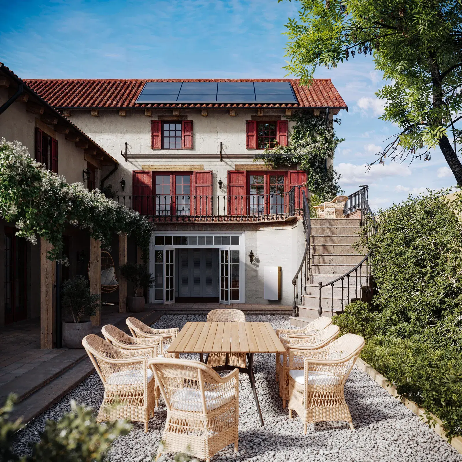 A row of dark rectangular solar panels mounted on the tiled roof of a rustic two-story home, with a wicker-furnished gravel patio in the foreground.