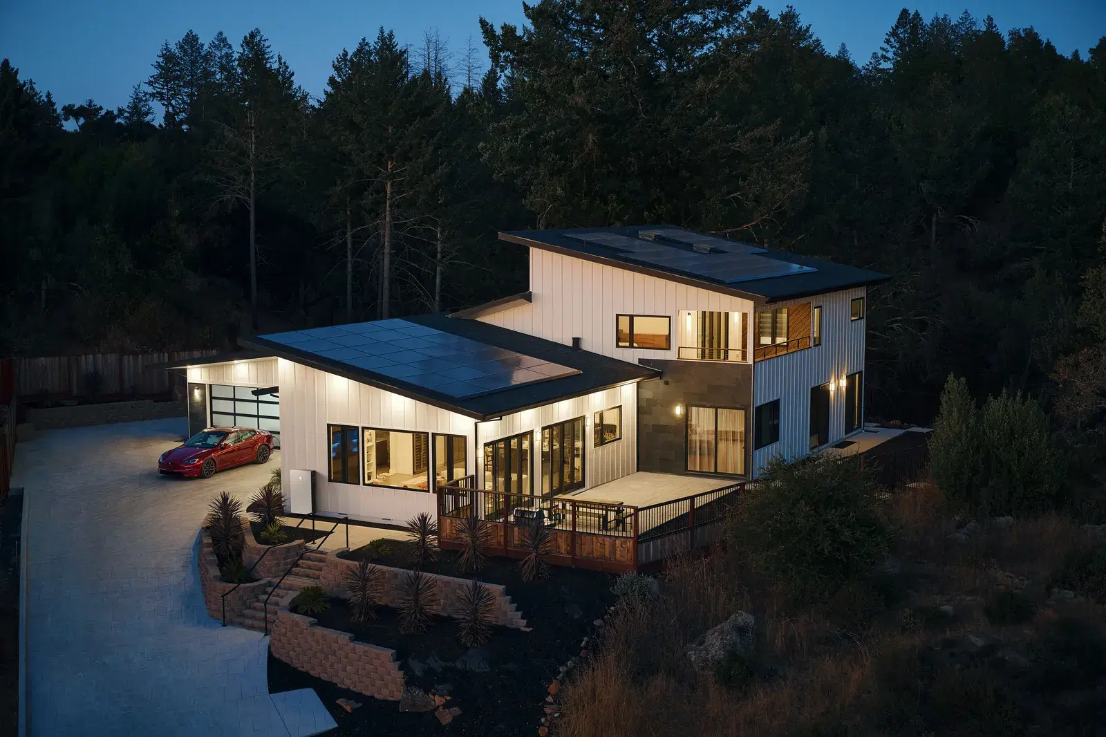 Aerial twilight view of a contemporary white home with rooftop solar panels and a red Tesla parked in the garage.