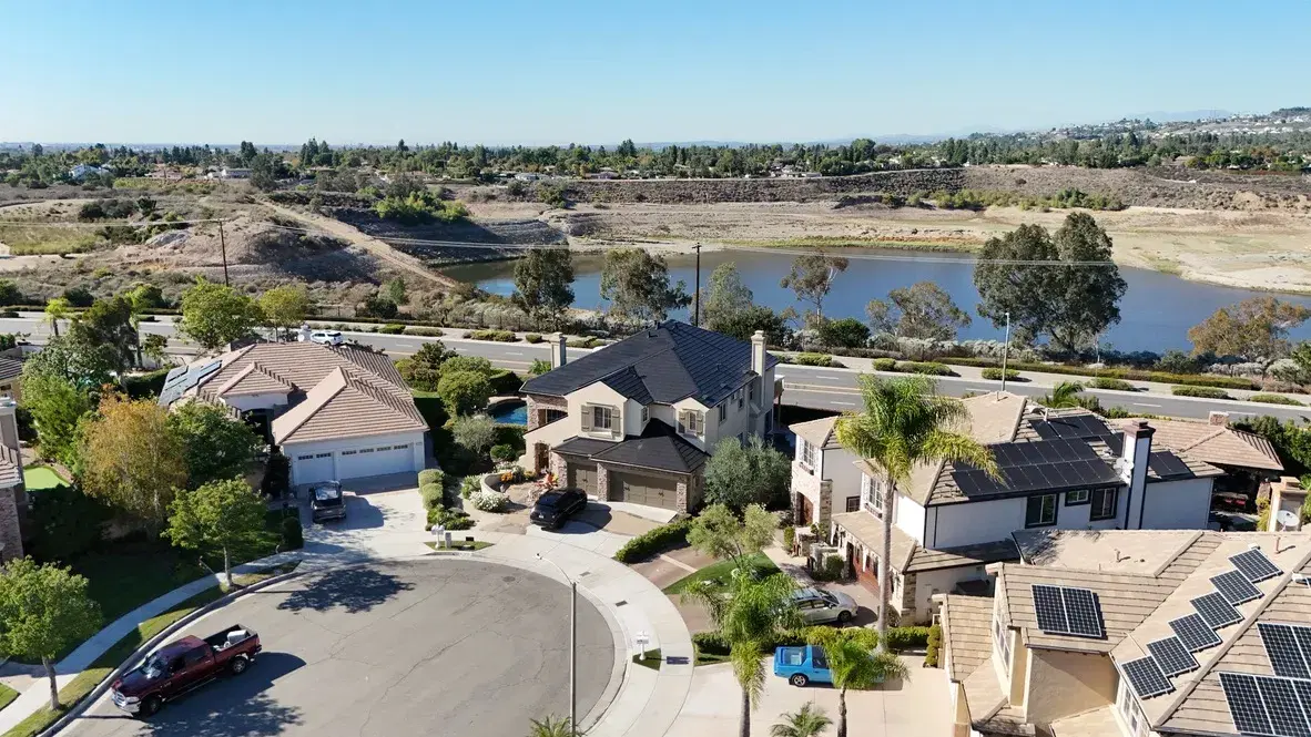 Aerial view of a suburban hillside neighborhood beside a reservoir, with rack-mounted solar panels on several home rooftops.