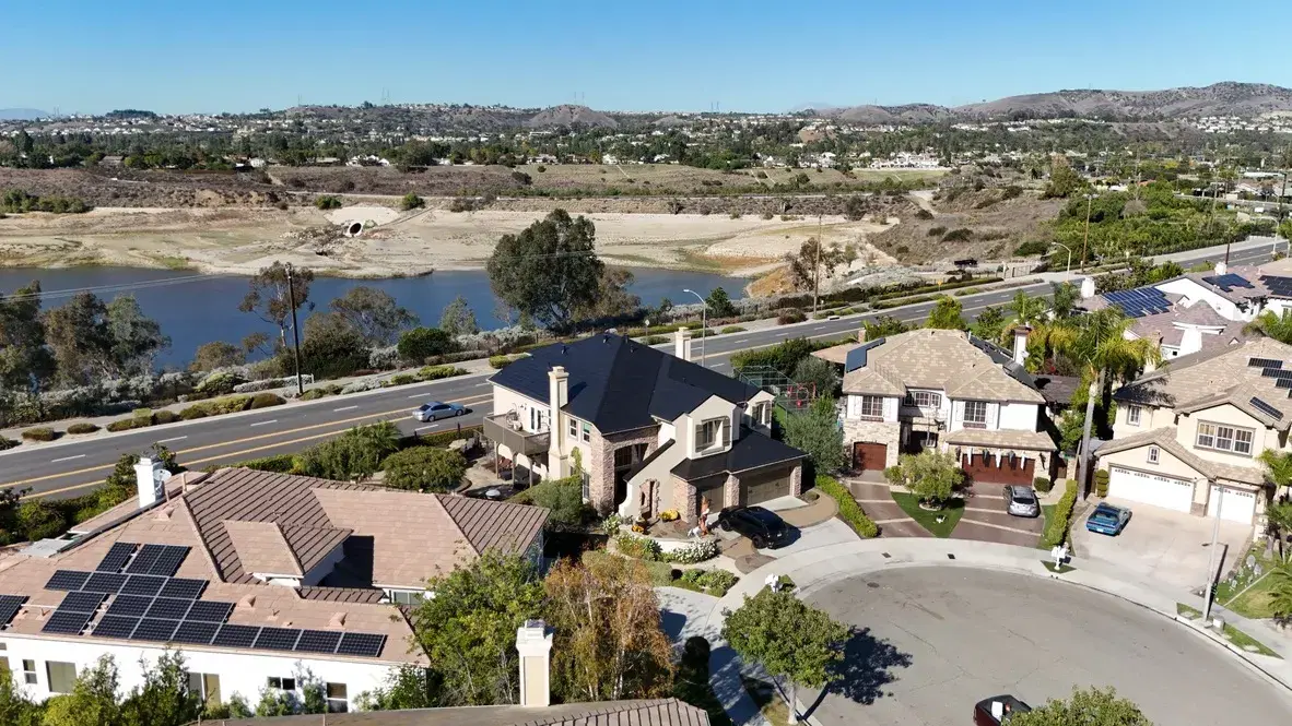 Aerial view of a suburban neighborhood near a reservoir with rack-mounted solar panel arrays on multiple rooftops and rolling hills beyond.