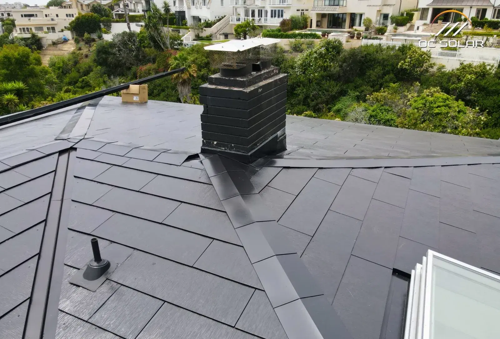Rooftop view of a completed Tesla Solar Roof made of smooth dark glass solar shingles, with a flashed chimney and skylight, overlooking a green hillside neighborhood.