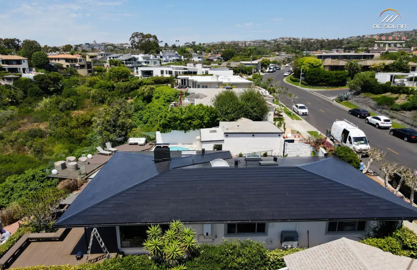Drone shot of a single-story home with a smooth dark glass solar-tile roof, surrounded by greenery and other houses on a hillside street.