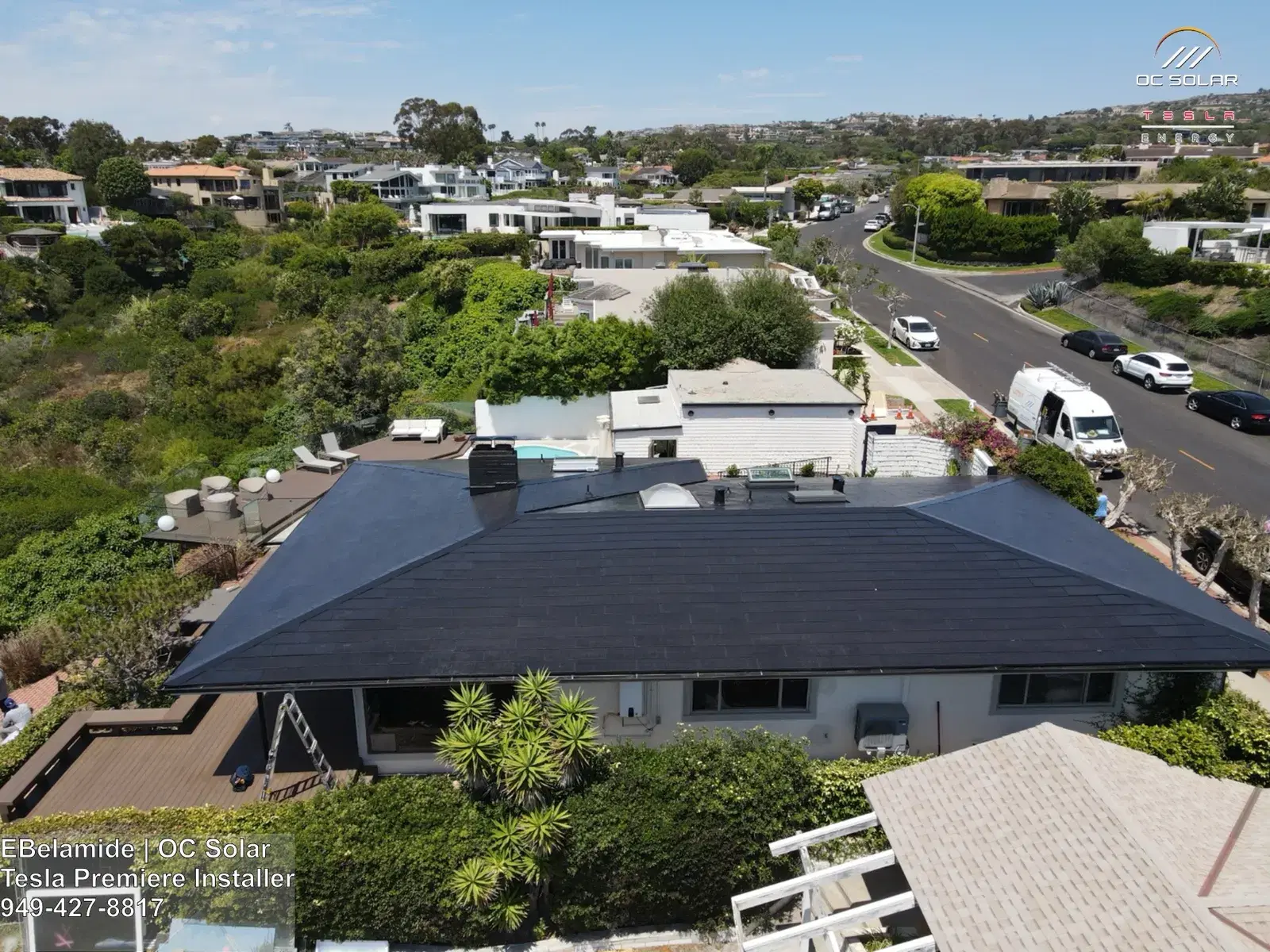 Drone shot looking down on a single-story home with a uniform dark Tesla Solar Roof, surrounded by greenery and ocean-view houses with a street and van below.