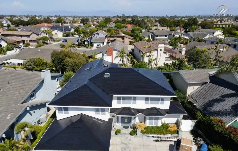 Drone photo looking down on a white two-story house topped entirely by a smooth dark Tesla Solar Roof, surrounded by a suburban neighborhood.