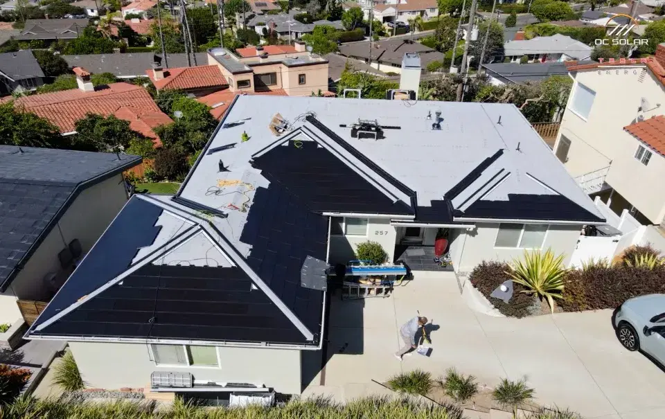 Drone view of a home with dark Tesla Solar Roof glass tiles covering most of the roof, materials and a worker visible below.