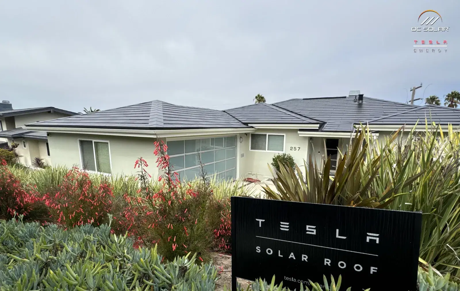 Single-story home with a dark glass Tesla Solar Roof and a black Tesla Solar Roof sign in the foreground garden.