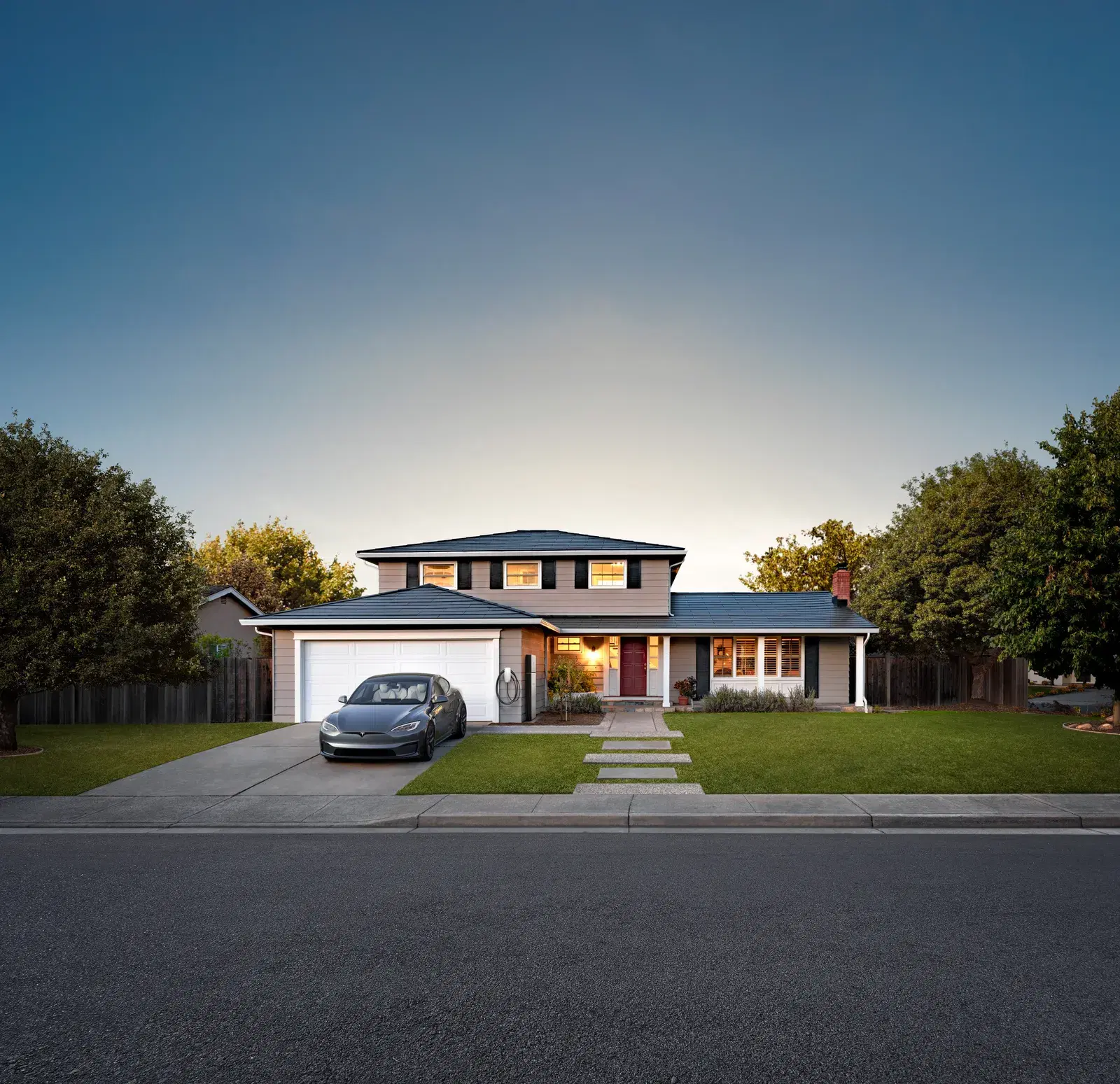 Dusk exterior of a two-story house with a smooth dark glass Tesla Solar Roof, warm interior lights glowing, and a Tesla in the driveway by a wall connector.