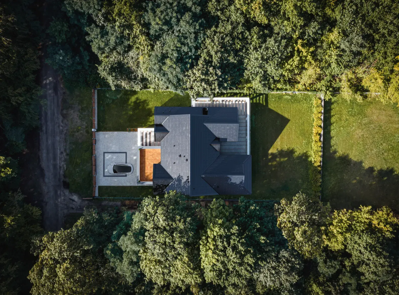 Overhead aerial of a house with a continuous dark glass Tesla Solar Roof surrounded by green woodland and lawn.