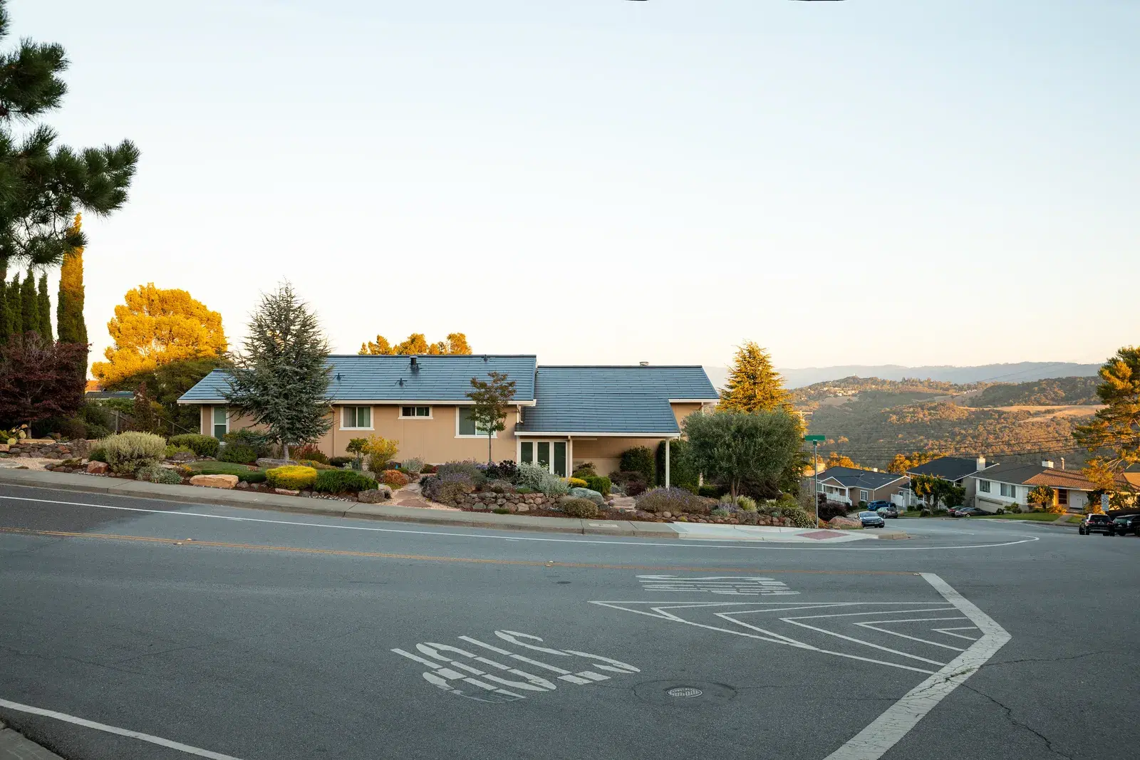 Single-story hillside home at sunset with a smooth dark glass Tesla Solar Roof, overlooking rolling hills and a wide street.