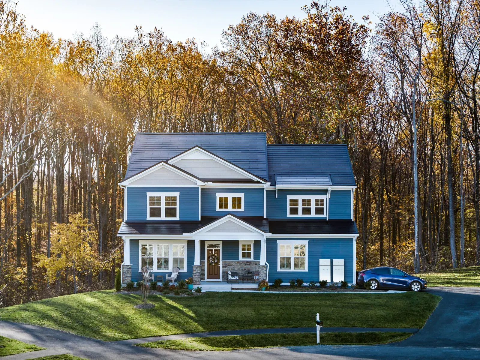 Blue craftsman two-story house with a dark glass Tesla Solar Roof, surrounded by fall trees.