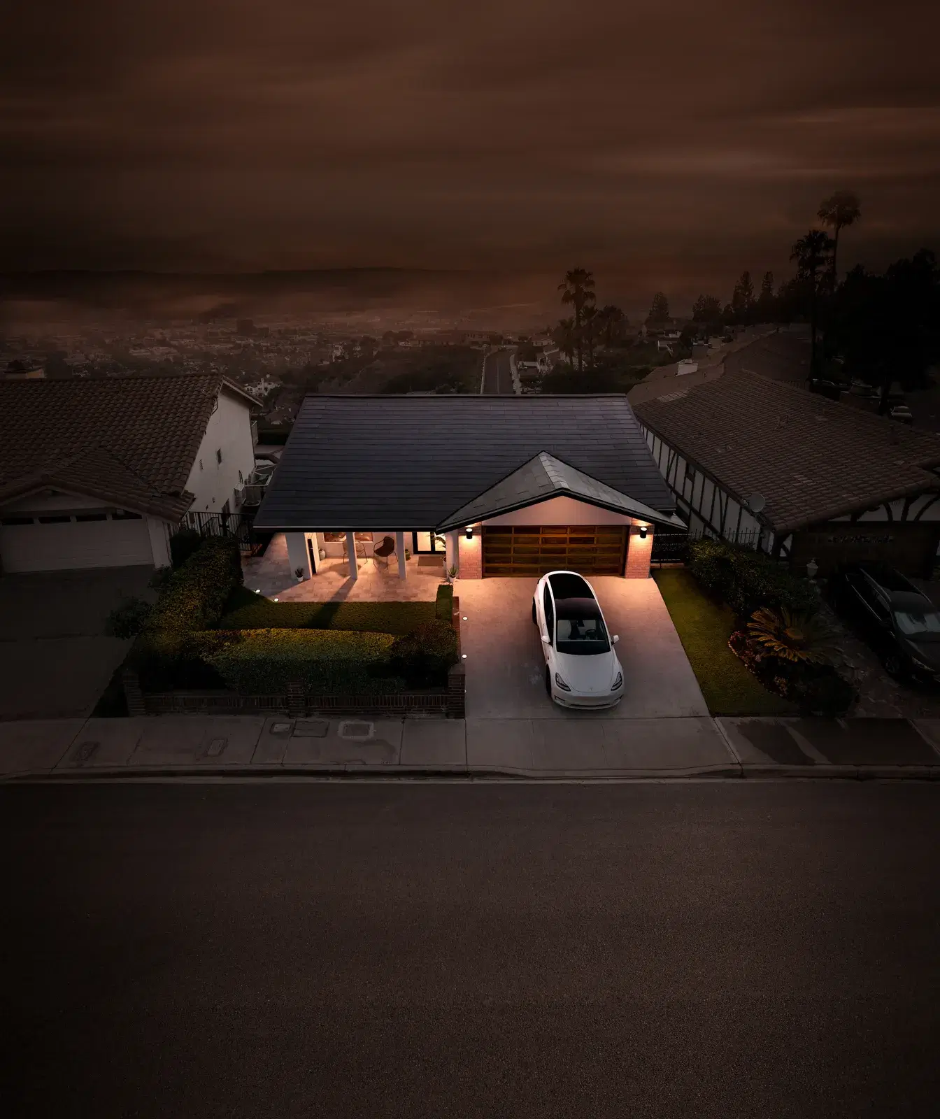 Aerial dusk view of a single-story home with a Tesla Solar Roof, lit from within, above a darkening cityscape.
