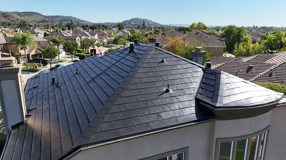 Drone view of a large home with a dark glass Tesla Solar Roof, with tile-roofed houses and hills behind.