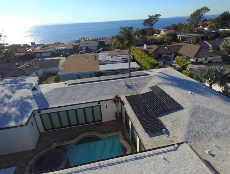 Drone view of an all-black OC Solar panel array on a coastal Southern California home, courtyard pool below and the Pacific Ocean on the horizon