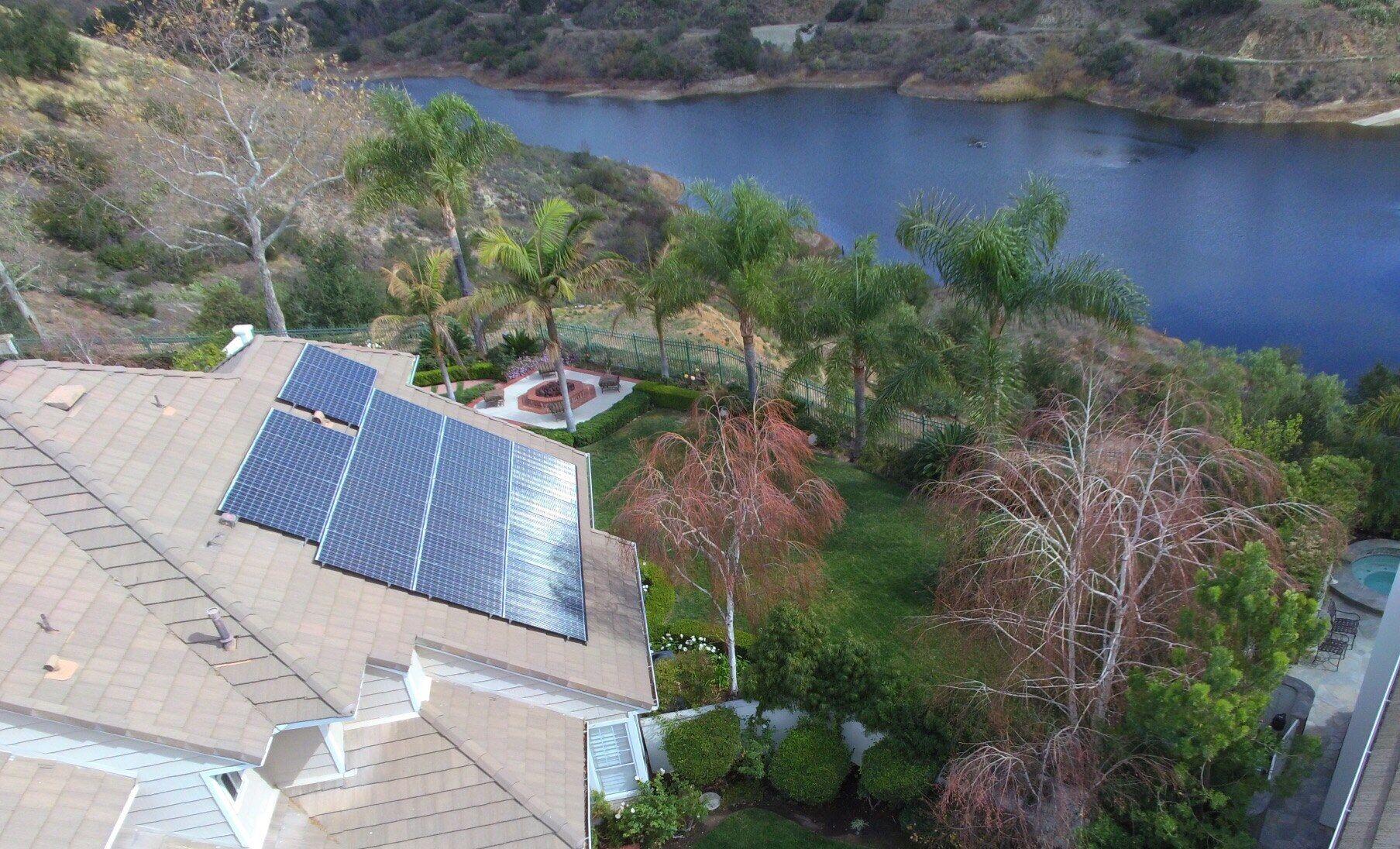 Drone view of an OC Solar rooftop array on a lakeside Southern California home, palm-lined shore behind