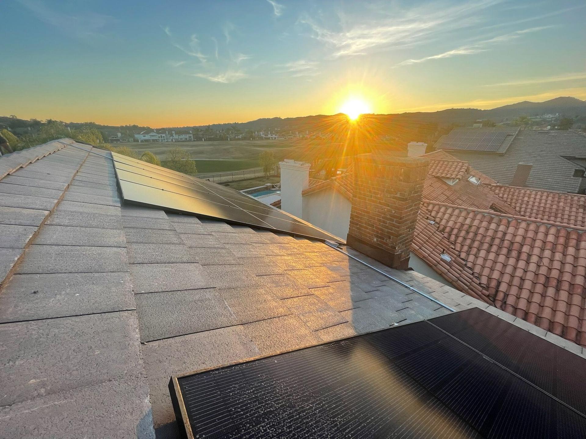 Golden-hour sunburst over an OC Solar rooftop array, terracotta roofs and hills beyond — installer's-eye view