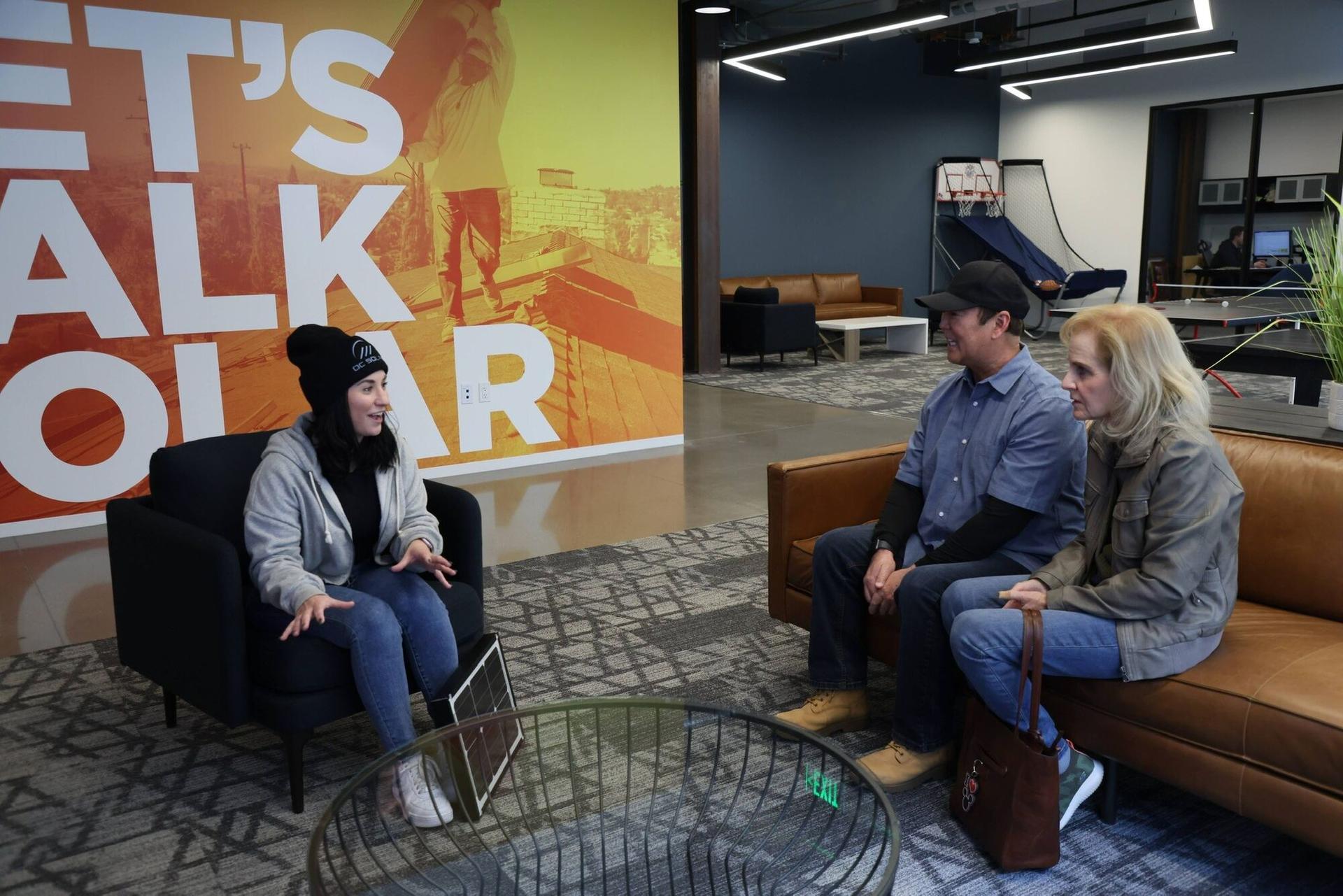 An OC Solar advisor sits down with a homeowner couple on the leather couches at the Irvine office, beside the orange 'Let's Talk Solar' wall mural
