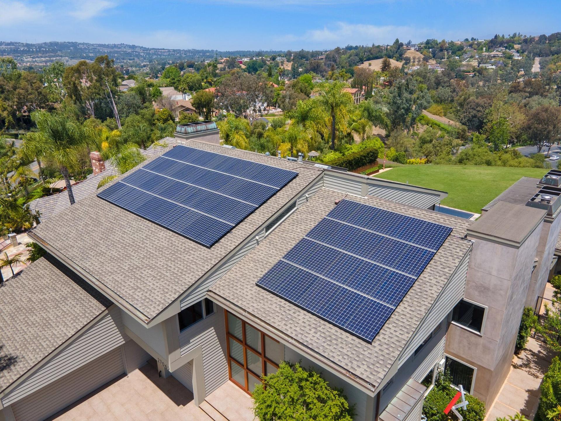 Drone view of two OC Solar arrays on a light-gray shingle roof with a skylight between them, green hills and neighboring homes beyond