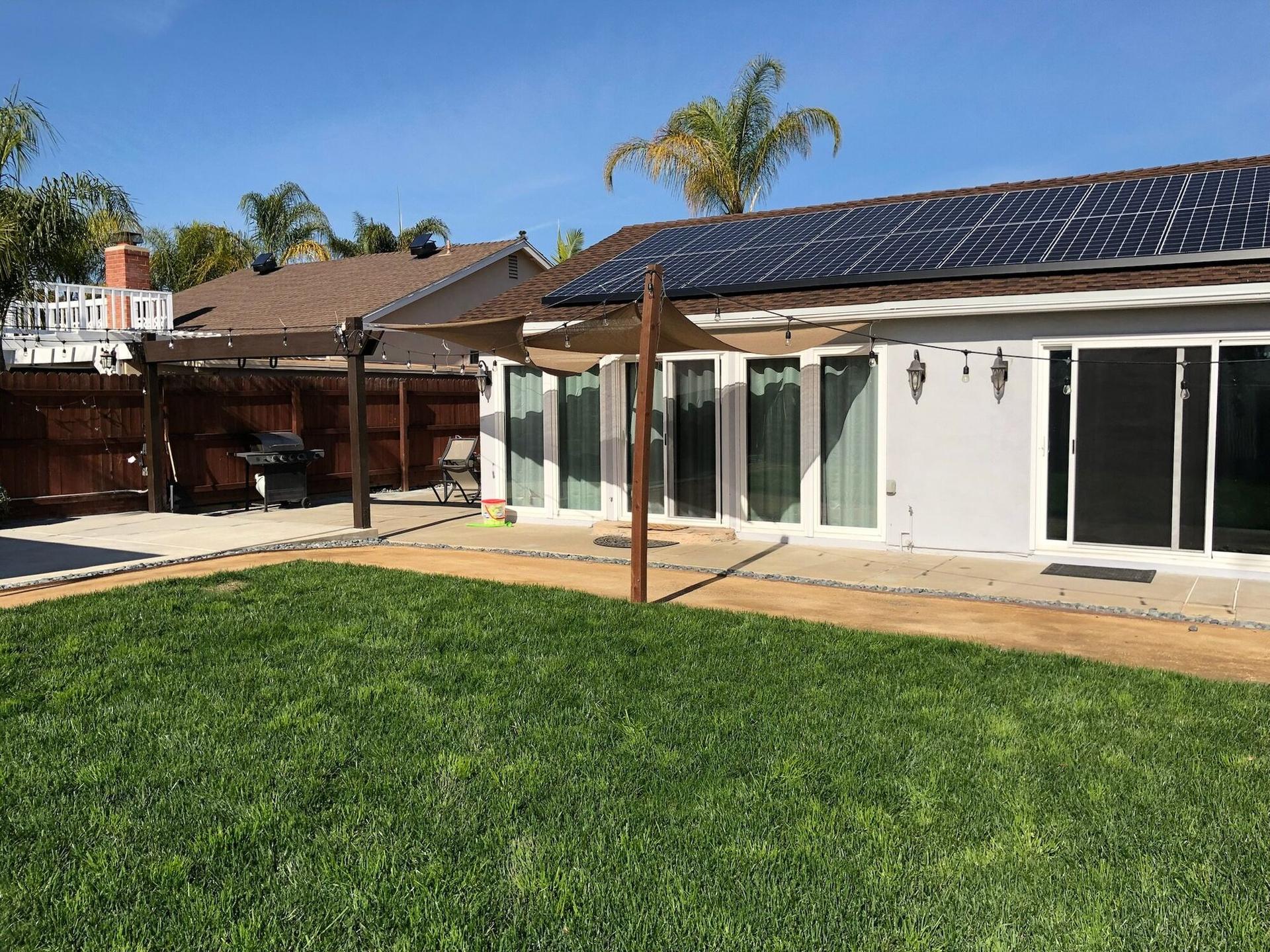 Backyard of a white single-story Southern California home with an all-black solar array on its brown shingle roof, green lawn, wood pergola with string lights, and shade sails over the patio