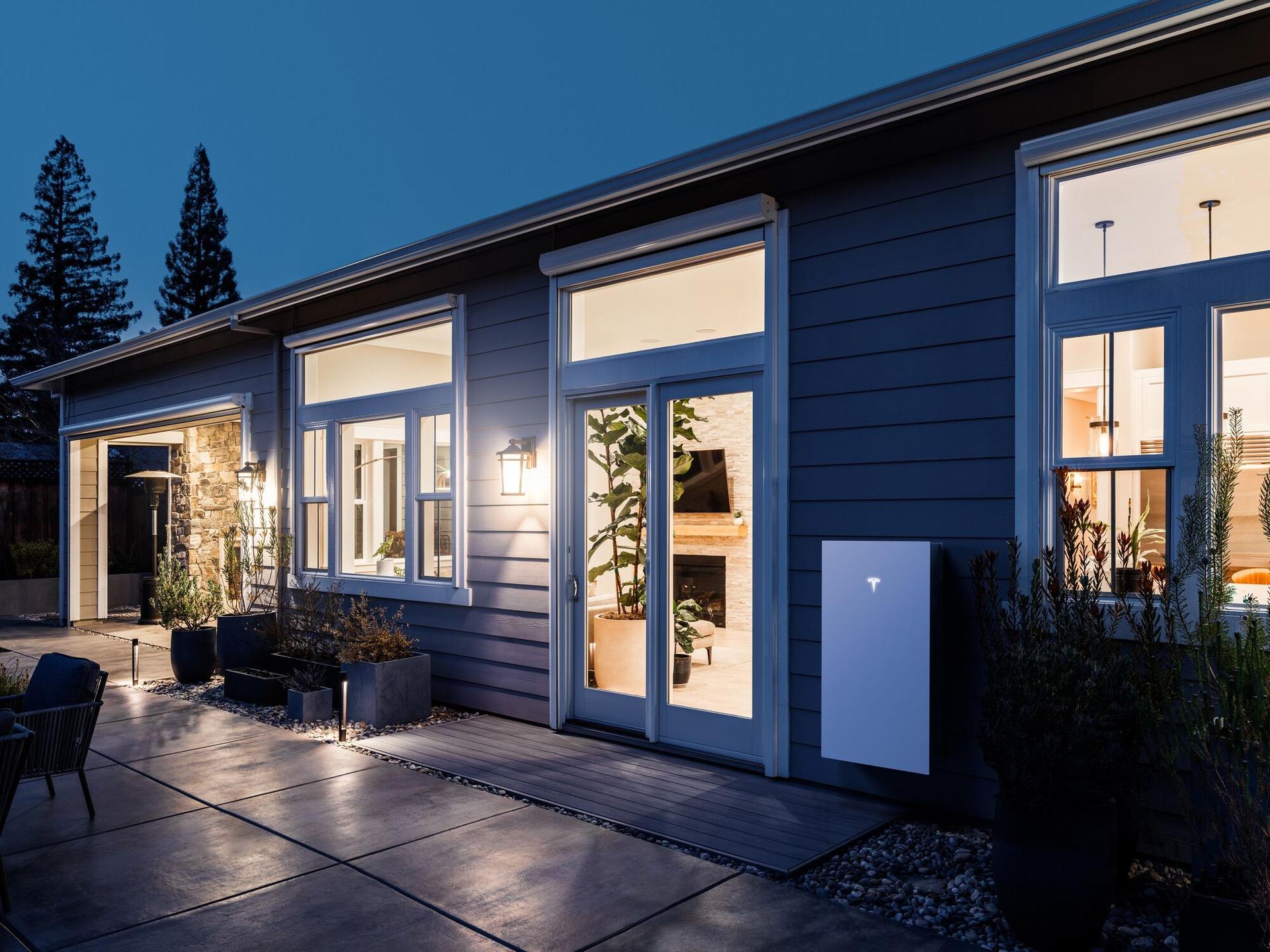Official Tesla render: a home at night with a wall-mounted Powerwall 3, lit windows, and patio