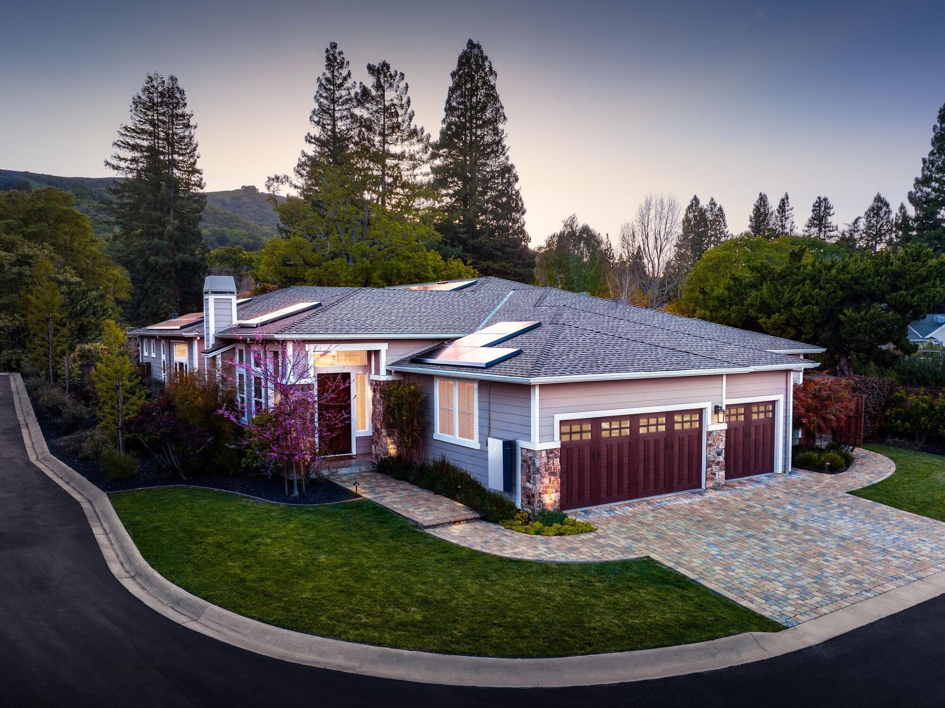 Official Tesla aerial dusk photo: an estate home with two rooftop solar arrays, lit entry windows, and garage sconces