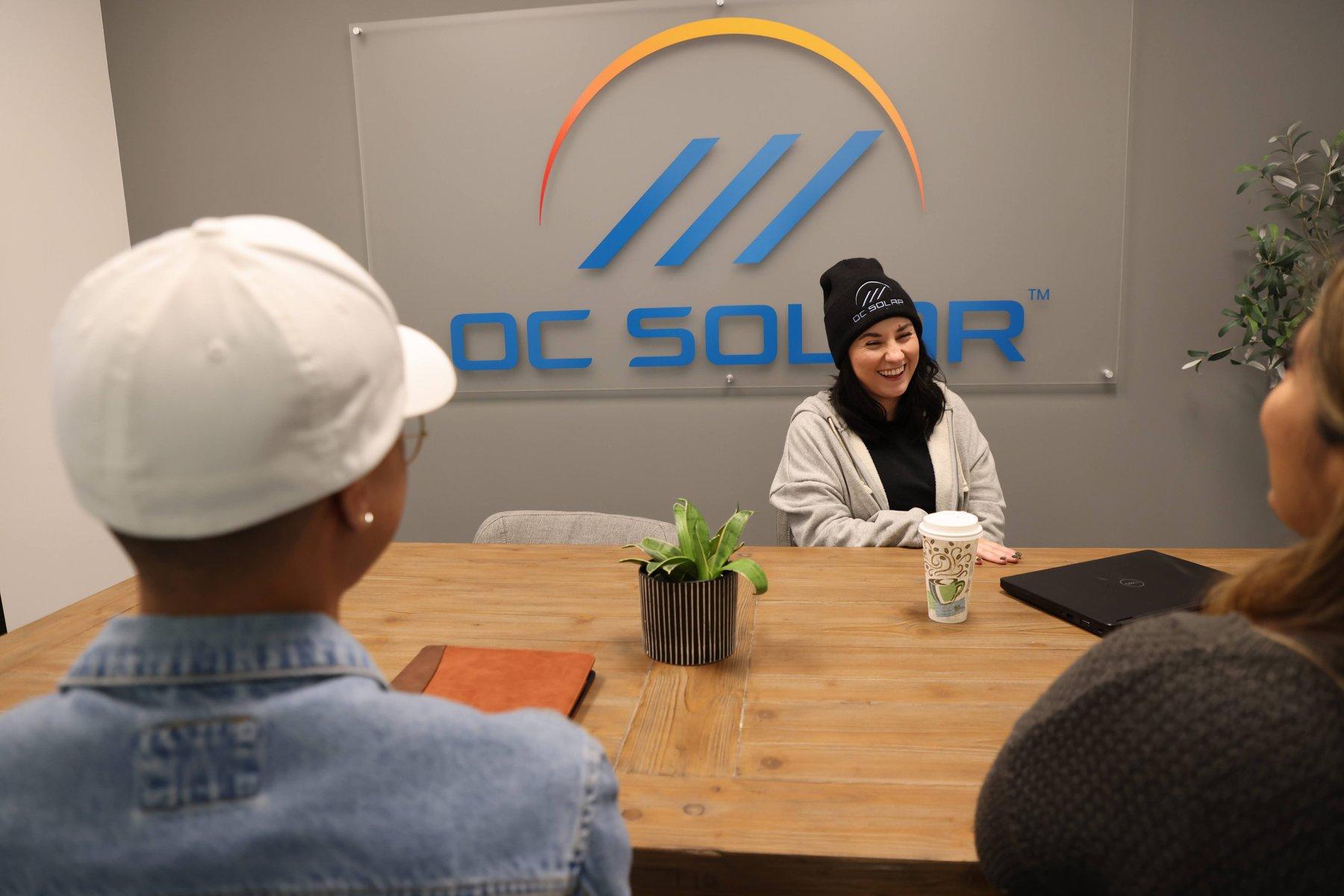 An OC Solar consultant in a branded beanie meeting two homeowners across a table beneath the OC Solar logo