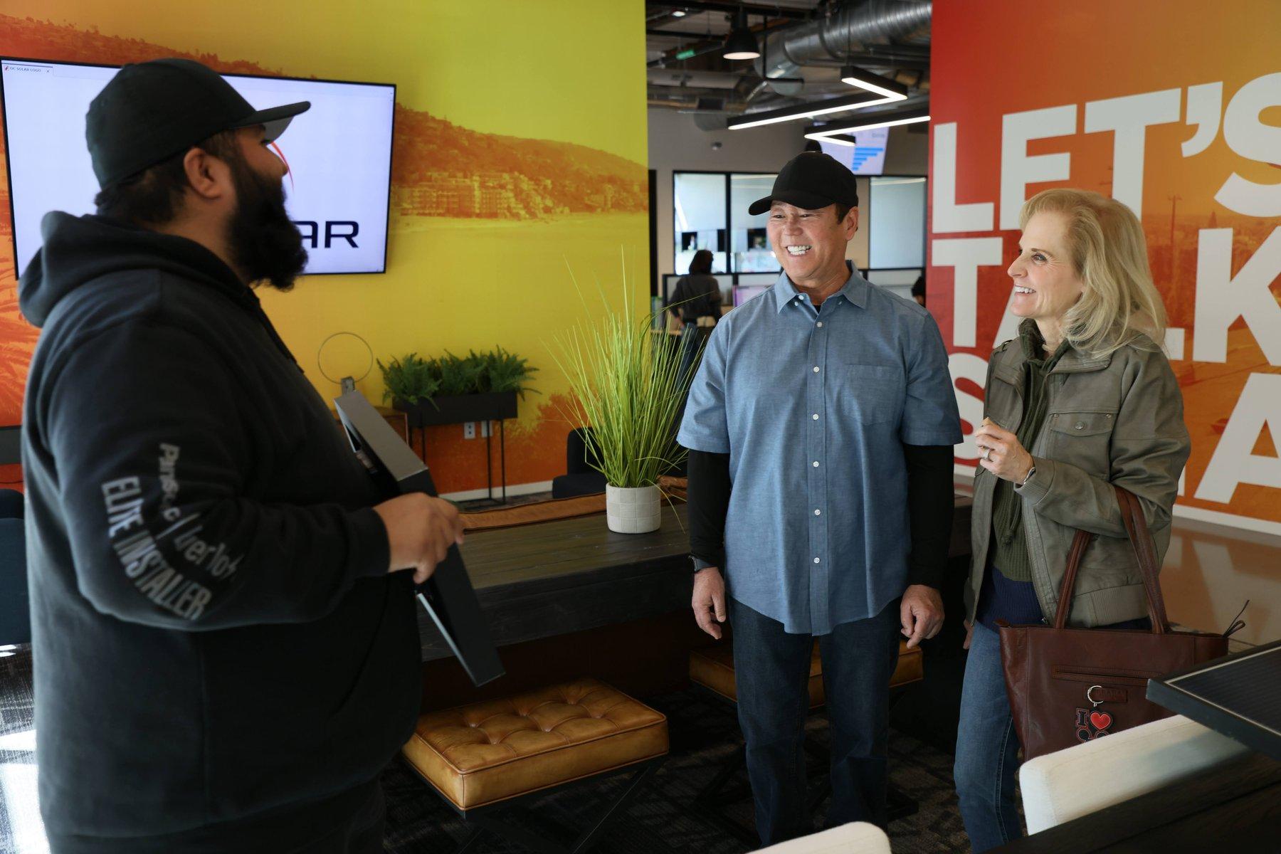 An OC Solar team member showing a solar panel to two smiling homeowners by the Let's Talk Solar wall in the Irvine office