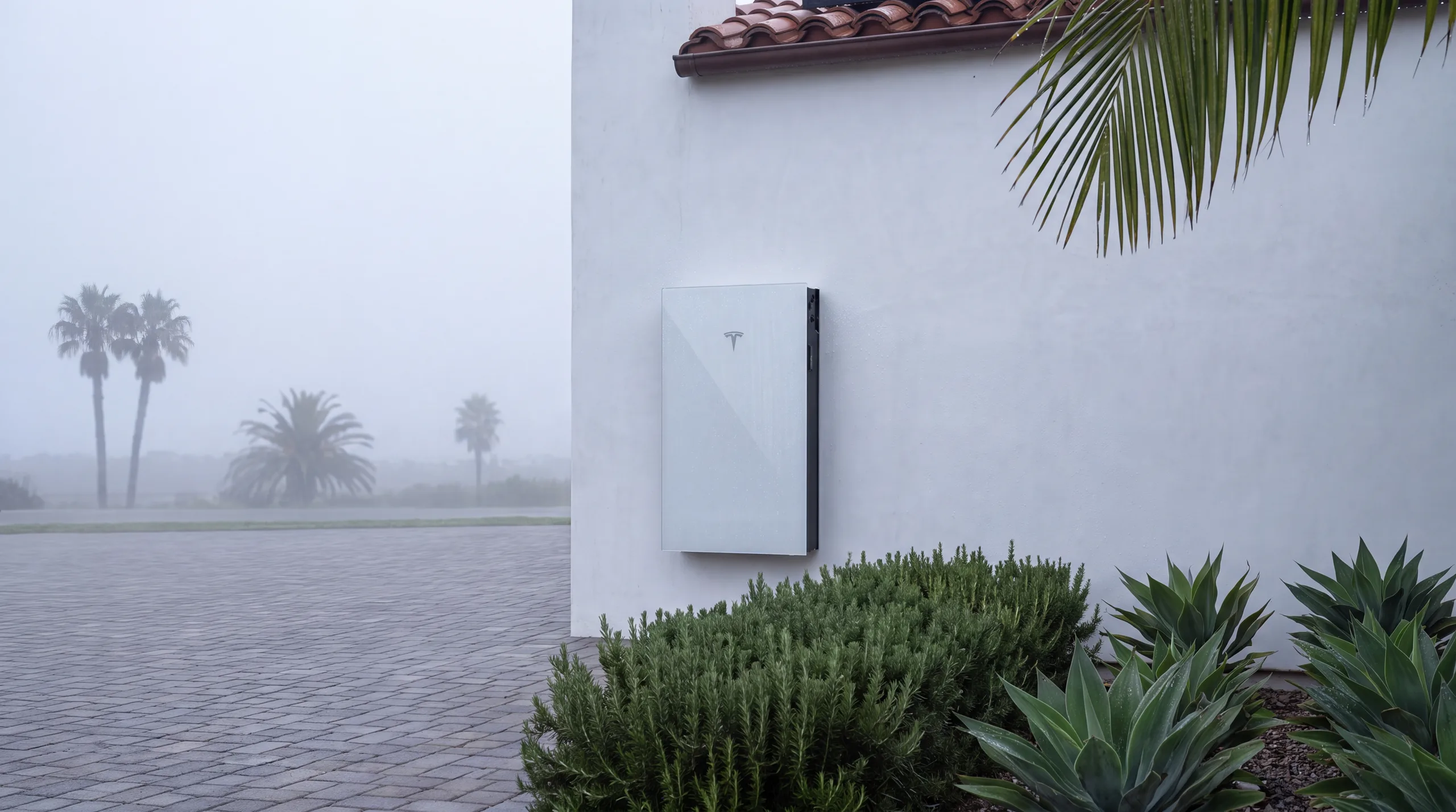 A Tesla Powerwall 3 mounted flush on the white stucco side wall of a Southern California home in marine-layer dawn light